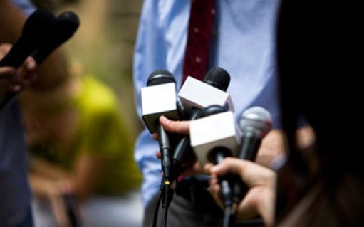 man in blue shirt being interviewed by media with several microphones